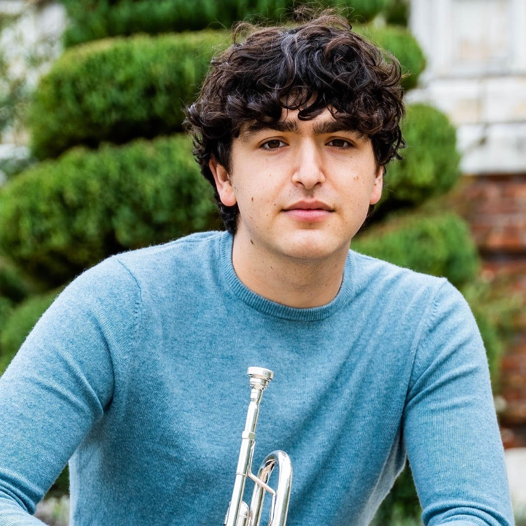 A photo of Jake wearing a blue sweater holding a trumpet.